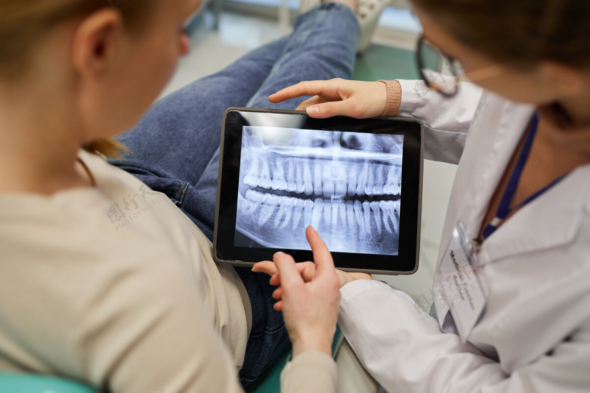 患者医生在牙科诊所向患者展示数字平板电脑上的牙齿x光片的后视图室内职业特写