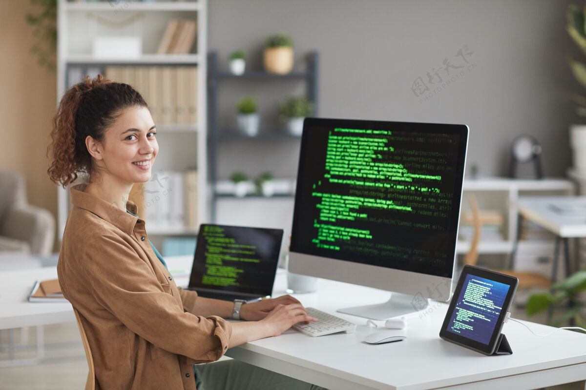成功年轻女子坐在电脑显示器前的桌子旁 一边用软件工作 一边在前面微笑的画像开发互联网成人