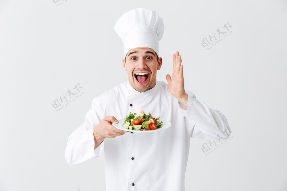 午餐兴奋的男厨师穿着制服 把新鲜的蔬菜沙拉放在隔离在白色墙上的盘子上成功专业烹饪