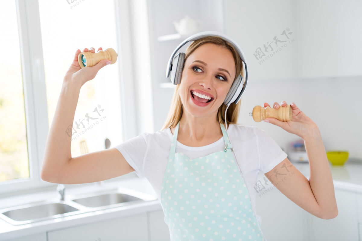 盐美丽的家庭主妇花上午听音乐的照片凉爽的现代耳机耳朵享受旋律拿着胡椒容器白色厨房室内唱歌围裙耳机