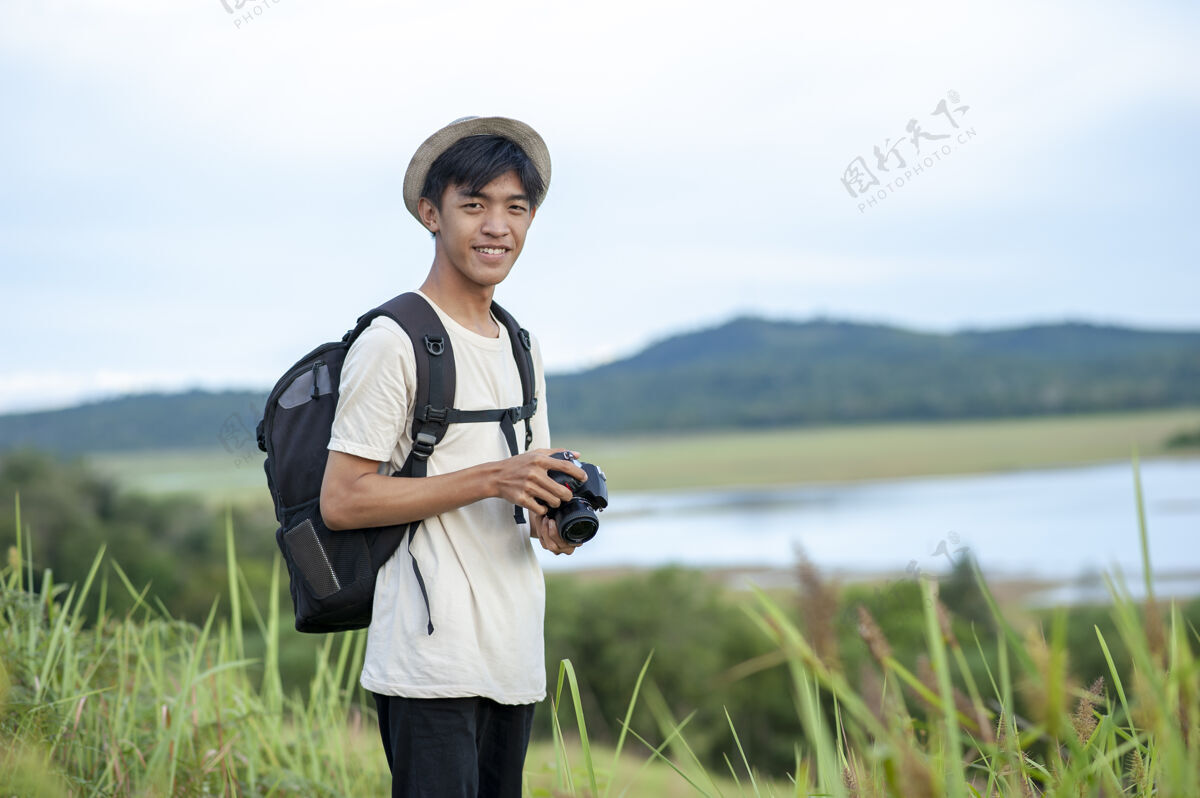 坐骑年轻的亚洲男人旅行者背着背包 用数码相机看着相机美丽的自然湖背景男人旅行湖边