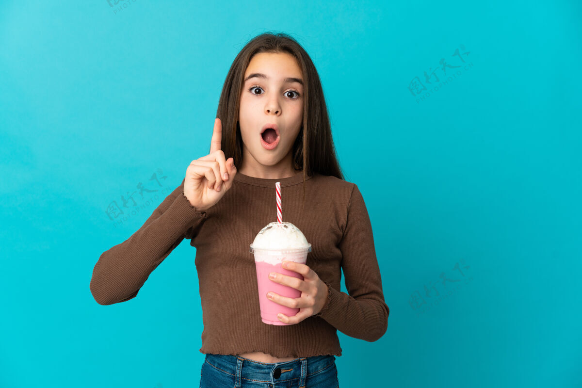 解决方案拿着草莓奶昔的小女孩被隔离在蓝色的背景上 她想在抬起手指的同时实现解决方案冰沙食物实现