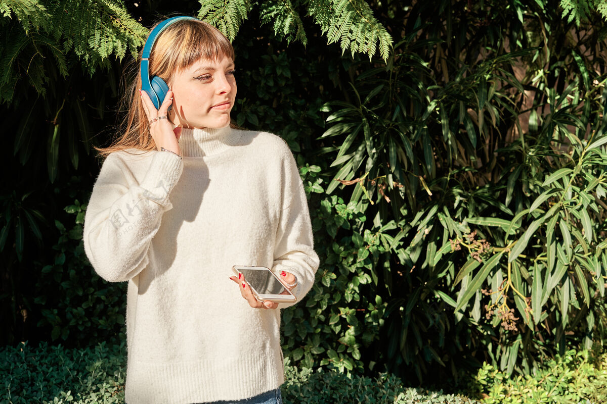 模型一个漂亮的白人女孩在公园里戴着蓝色耳机听音乐公园女孩姿势