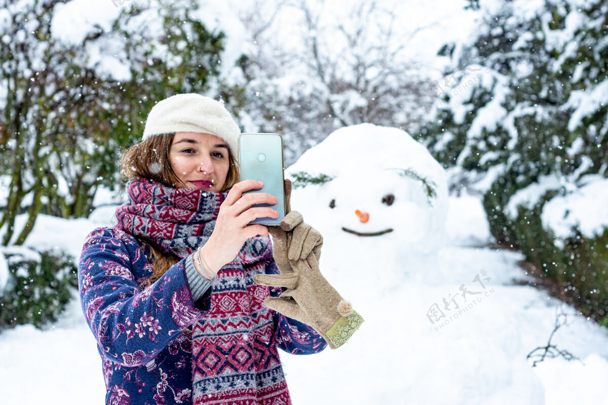 活动一个戴着贝雷帽 夹克和围巾的金发女人和雪人用手机自拍的照片雪花女人享受