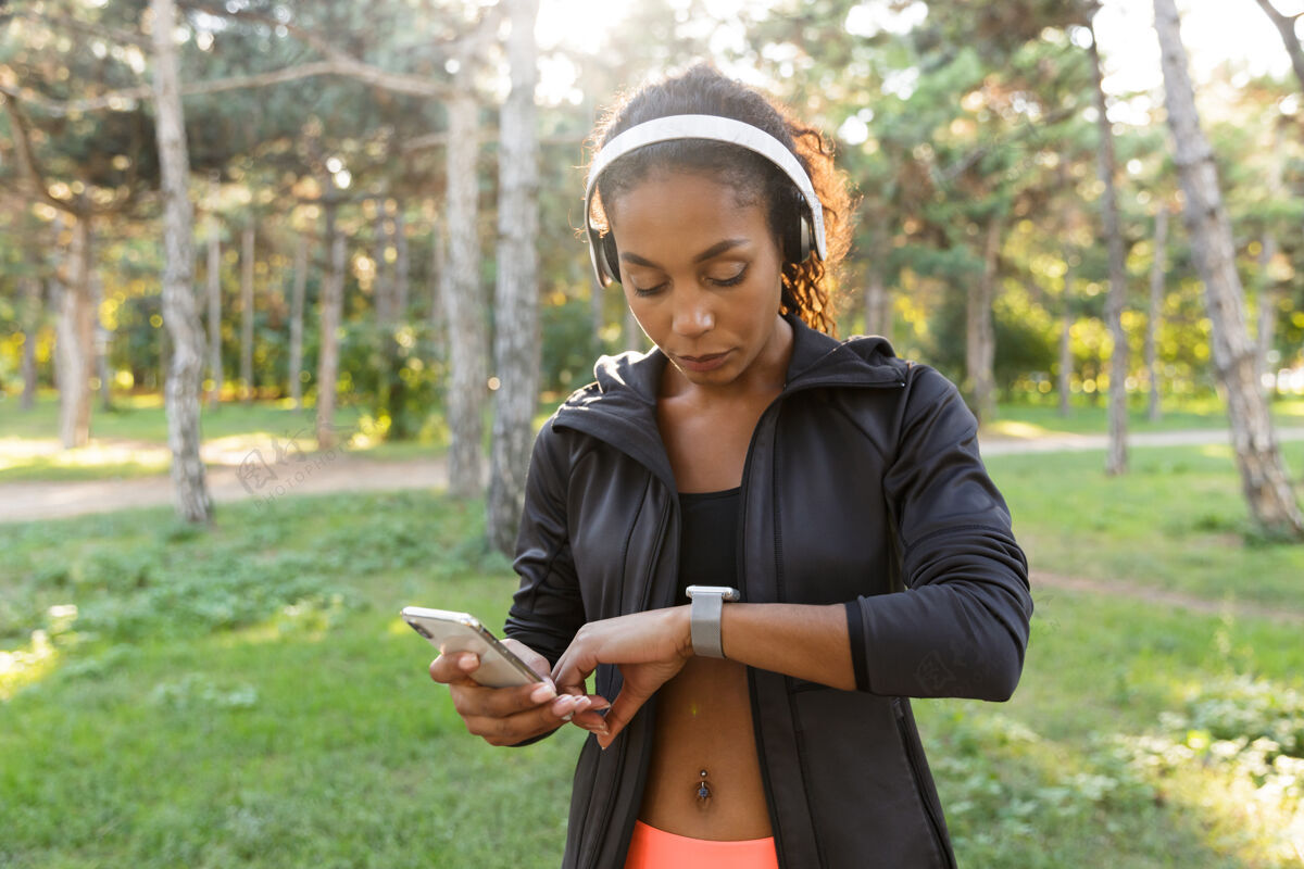 电话一张20多岁的健身女士的肖像 她穿着黑色运动服 戴着耳机 一边看着手表 一边穿过绿色公园身体成人运动