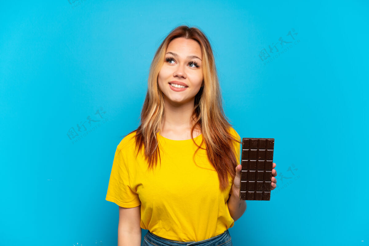 美味十几岁的女孩拿着巧克力在孤立的蓝色背景下思考一个想法 同时抬头想象聪明女人