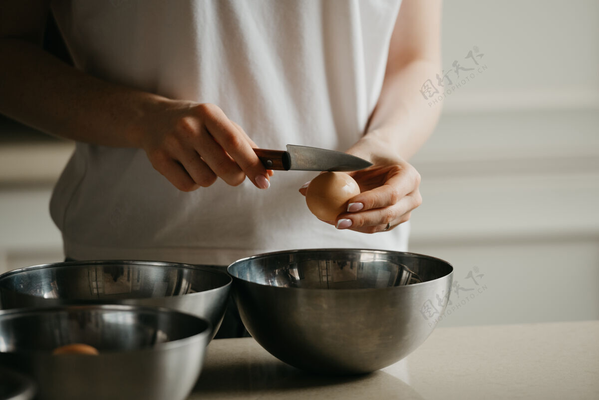 面包房厨房不锈钢碗上方 一名年轻女子用刀打碎蛋壳的照片烹饪准备鸡蛋
