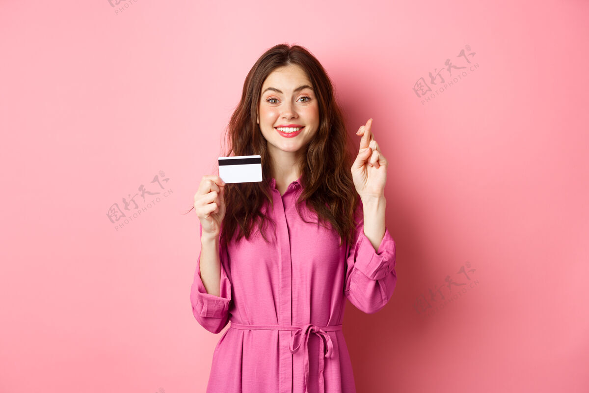 快乐一位充满希望的年轻女子 对着粉色的墙壁 展示着塑料信用卡和祈求好运的十指交叉 许下愿望 面带微笑候选人欢呼购物
