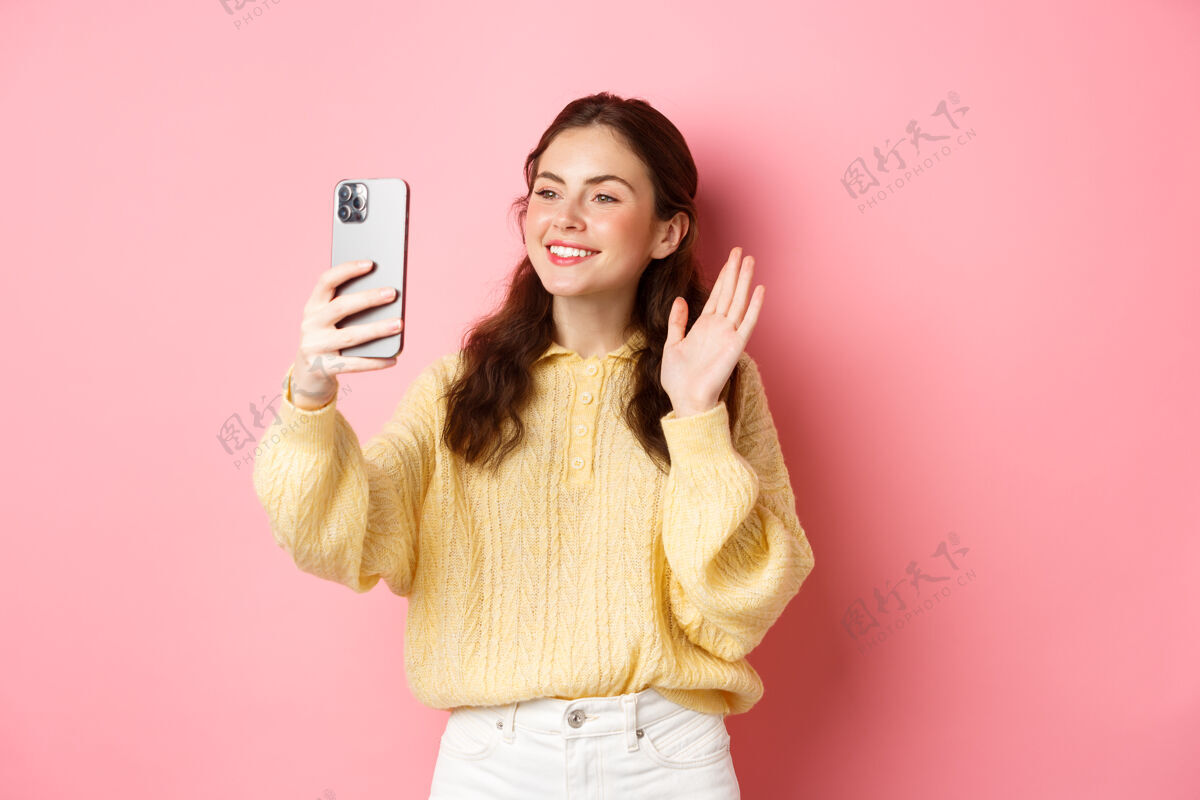 微笑在视频聊天应用程序上聊天的友好女孩的肖像 在智能手机摄像头前挥手 进行移动对话 站在粉红色的墙上应用程序积极年轻