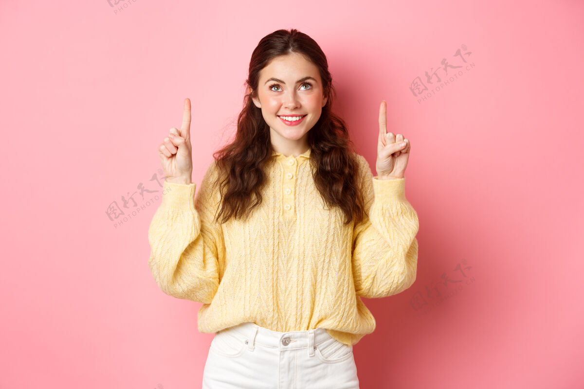 快乐美丽的年轻女子微笑的肖像 指着并仰望着顶级促销活动 展示广告 阅读促销文本 靠着粉色的墙壁站着手势欢呼学生