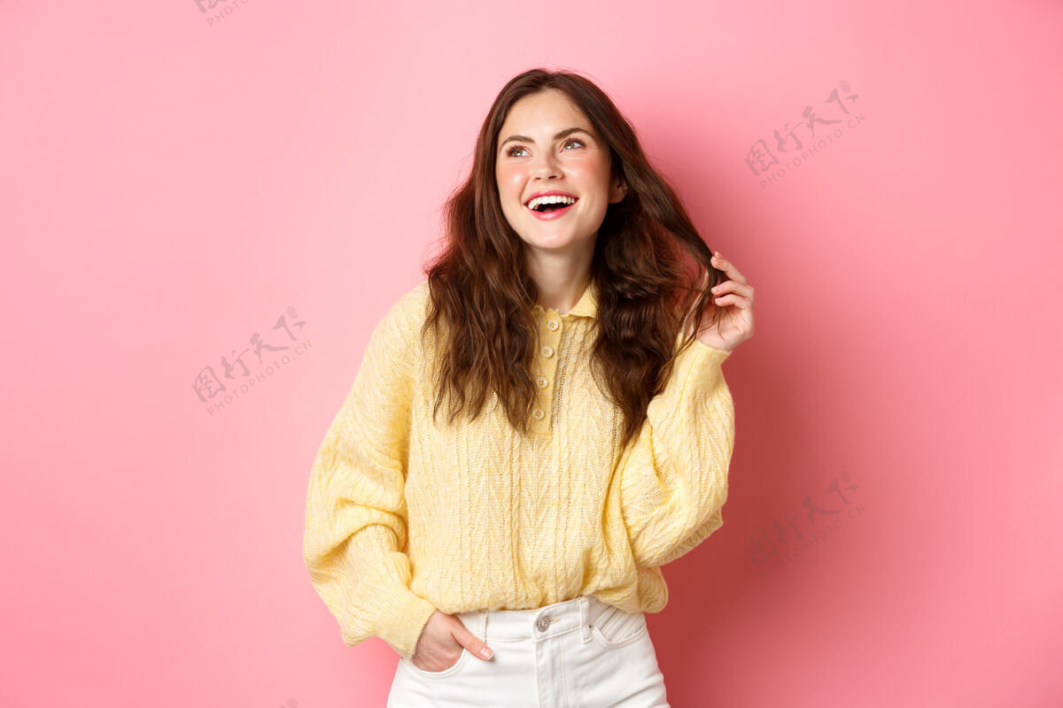 年轻20多岁的年轻女子一边笑一边玩着头发 一边看着左上角的宣传片 幸福地站在粉色的墙上黑发女孩学生