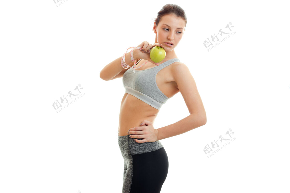 女士苗条的年轻女子侧身站着 低头 手里拿着隔离在白墙上的苹果锻炼健康磁带