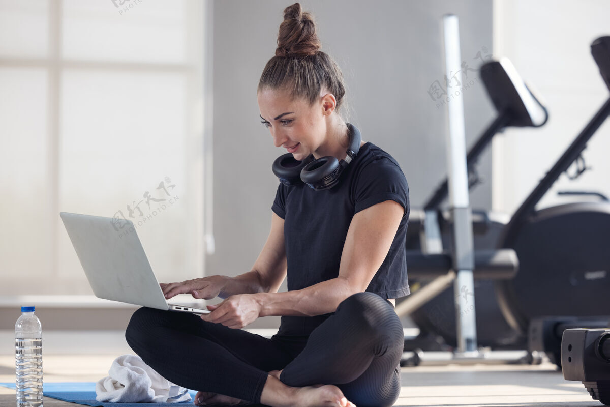 训练一个穿着运动服的运动型女人坐在地板上 手里拿着哑铃 蛋白奶昔或一瓶水 在家里的起居室里用着笔记本电脑房间.运动和娱乐理念健康生活方式瑜伽学习