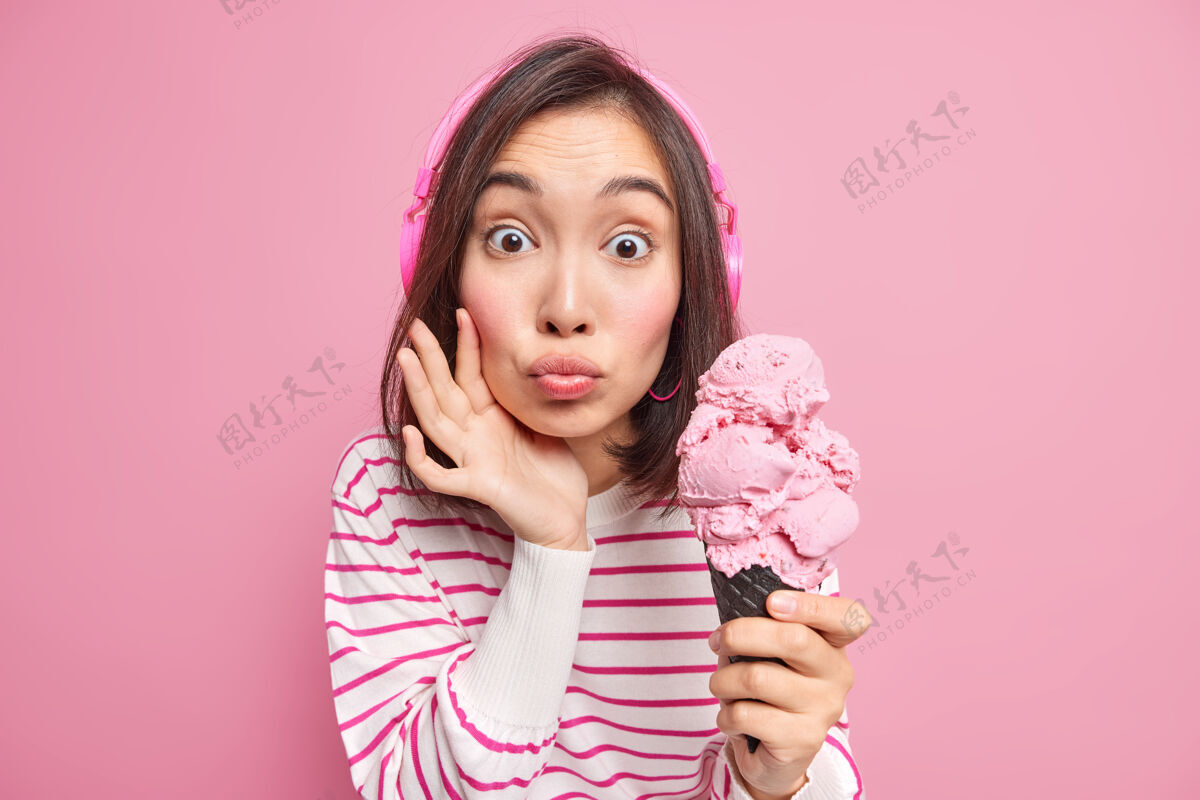 女性惊讶的亚洲女人嘴唇圆润吃着美味的冰淇淋想吃冰冻甜点耳朵上戴着耳机休闲条纹套头衫隔着粉色的墙壁夏天到了食物风味无线