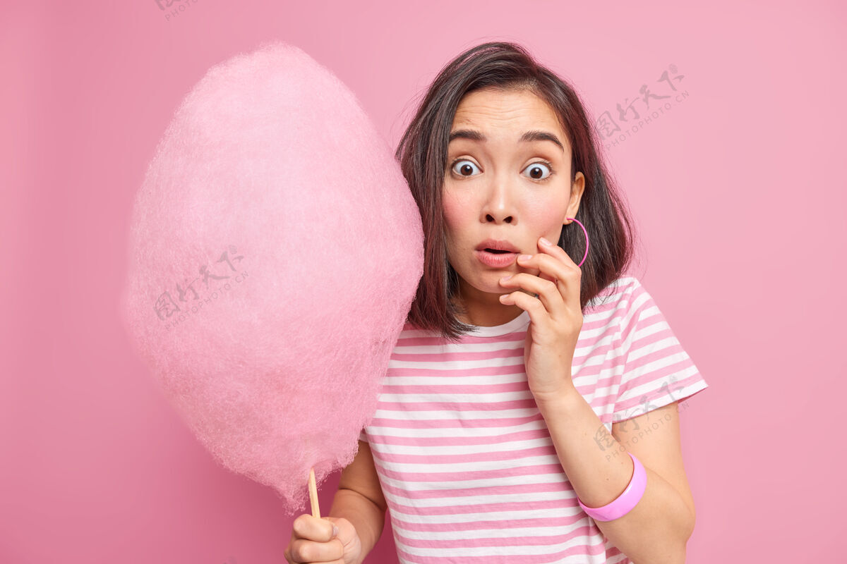 小吃吓得目瞪口呆的黑发女人拿着甜甜的棉花糖惊讶地听到一些惊人的发现她要吃多少卡路里穿条纹t恤隔离在粉红色的墙壁上糖果女人棉花