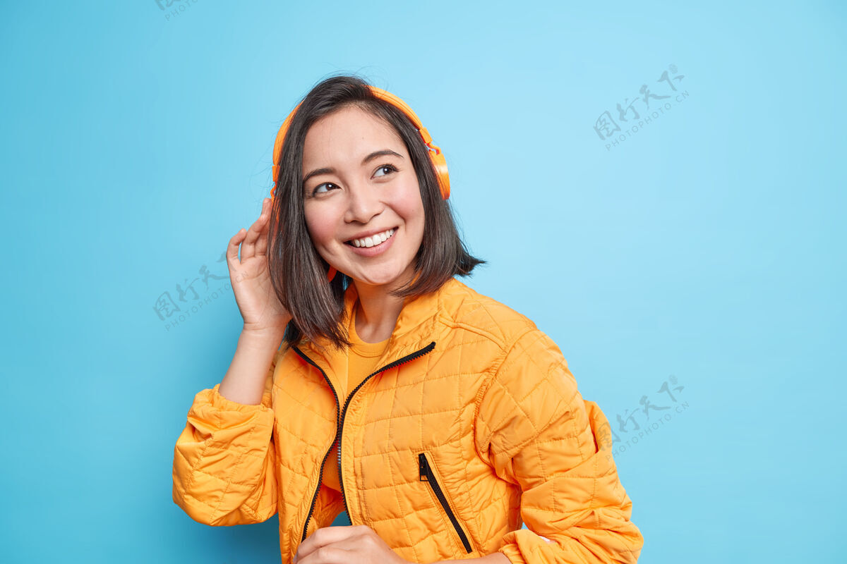 立体声一张漂亮的东方女孩的照片戴着现代的耳机听音乐微笑着宽大地穿着橙色的时尚夹克隔着蓝色的墙很棒的播放列表音乐爱好者转身穿微笑