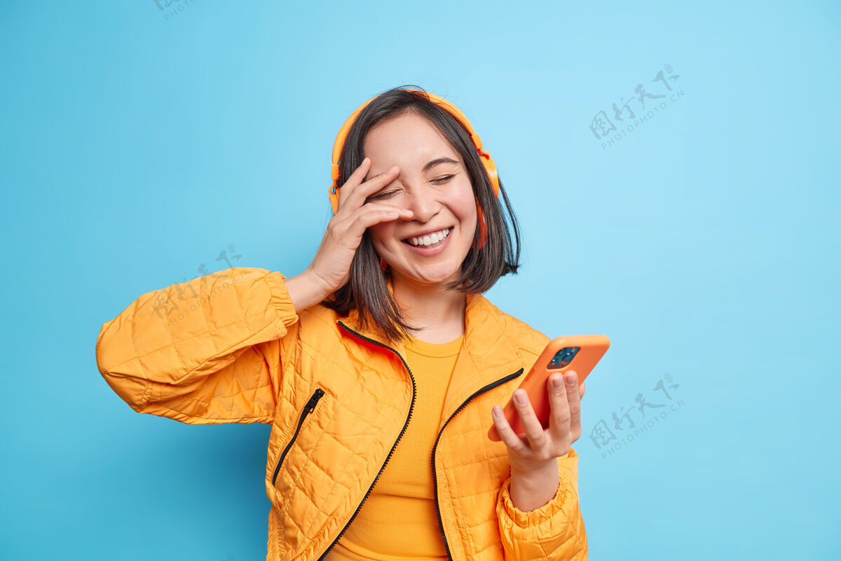 人物无忧无虑的时尚亚洲女人微笑积极闭上眼睛手放在脸上有乐趣听音乐手持现代智能手机戴无线耳机表达积极情绪摆出室内姿势乐趣电话穿着