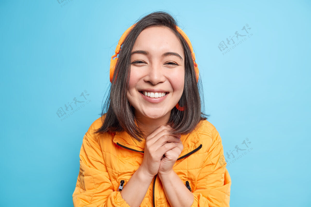 休闲快乐美丽的亚洲女人的画像双手合十微笑宽大享受良好的声音与现代耳机听音乐播放列表穿着橙色夹克隔离在蓝色的墙壁感情欢乐站