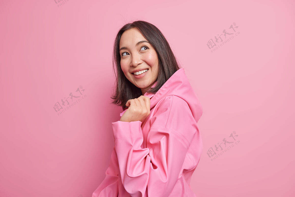微笑很高兴深色头发的亚洲女人站在粉红色的墙上 心情很好 穿着时髦的夹克 带兜帽 想着一些愉快的姿势 快乐的室内情感概念女性罗西漂亮