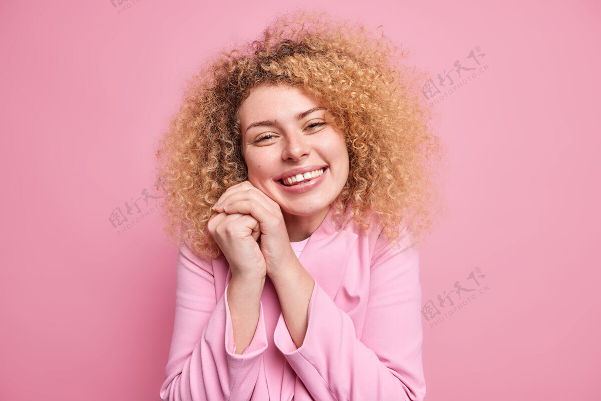 快乐快乐真诚的女人的肖像 卷发 手靠近脸 高兴地听到温暖的话语或恭维的微笑 身着粉色夹克 表达积极的情绪和感情温柔欢呼单色