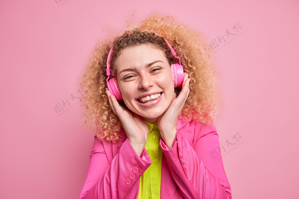 明亮积极的卷发少女喜欢听喜爱的音乐戴着立体声耳机心情愉快穿着鲜艳的衣服隔着粉色的墙壁快乐的女梅洛曼听歌曲耳机青少年积极