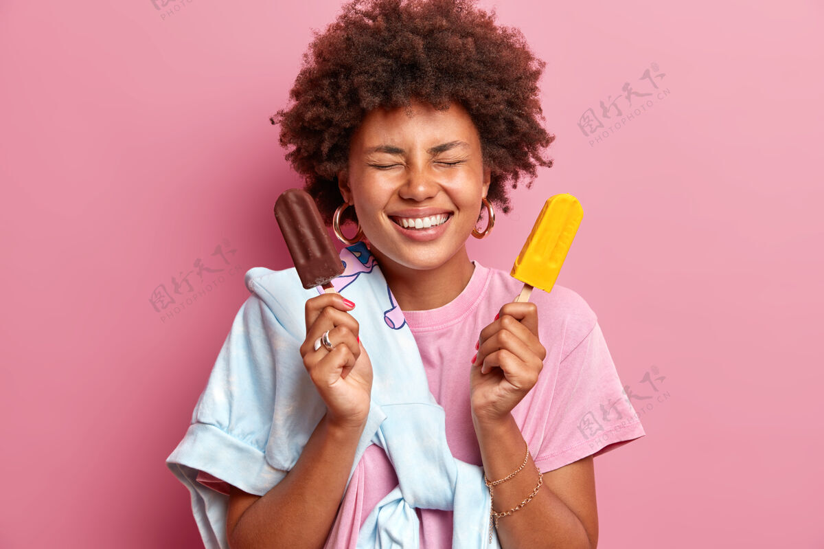 热量快乐的卷曲的美国黑人女人从快乐中咧嘴笑闭上眼睛摆出两个美味的冰激凌的姿势在粉色的墙上表达积极的情绪快乐的夏天冰青少年食物
