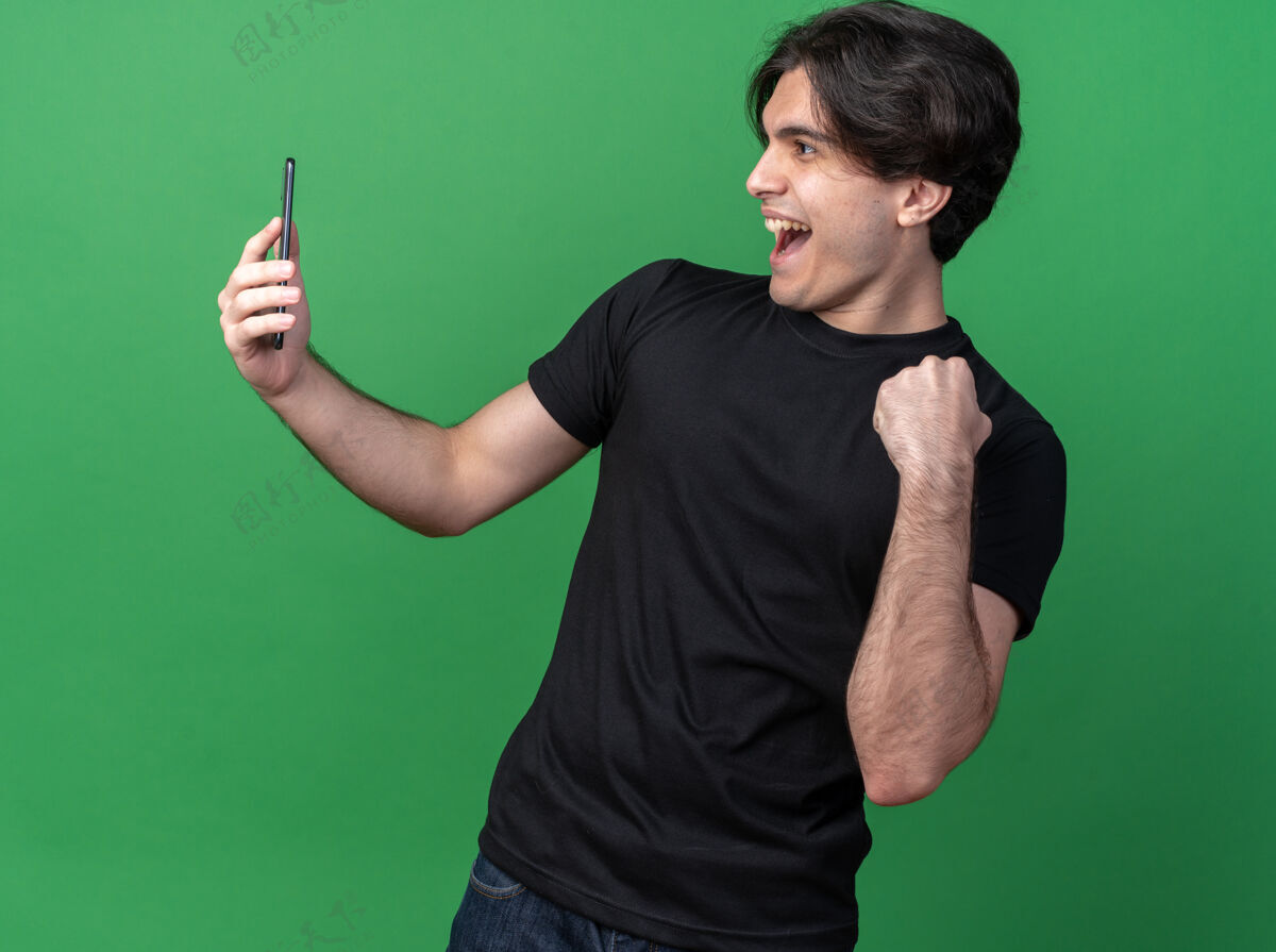 抱着快乐的年轻帅哥穿着黑色t恤 拿着手机 看着手机 在绿色的墙上显示“是”的手势穿着市民年轻人