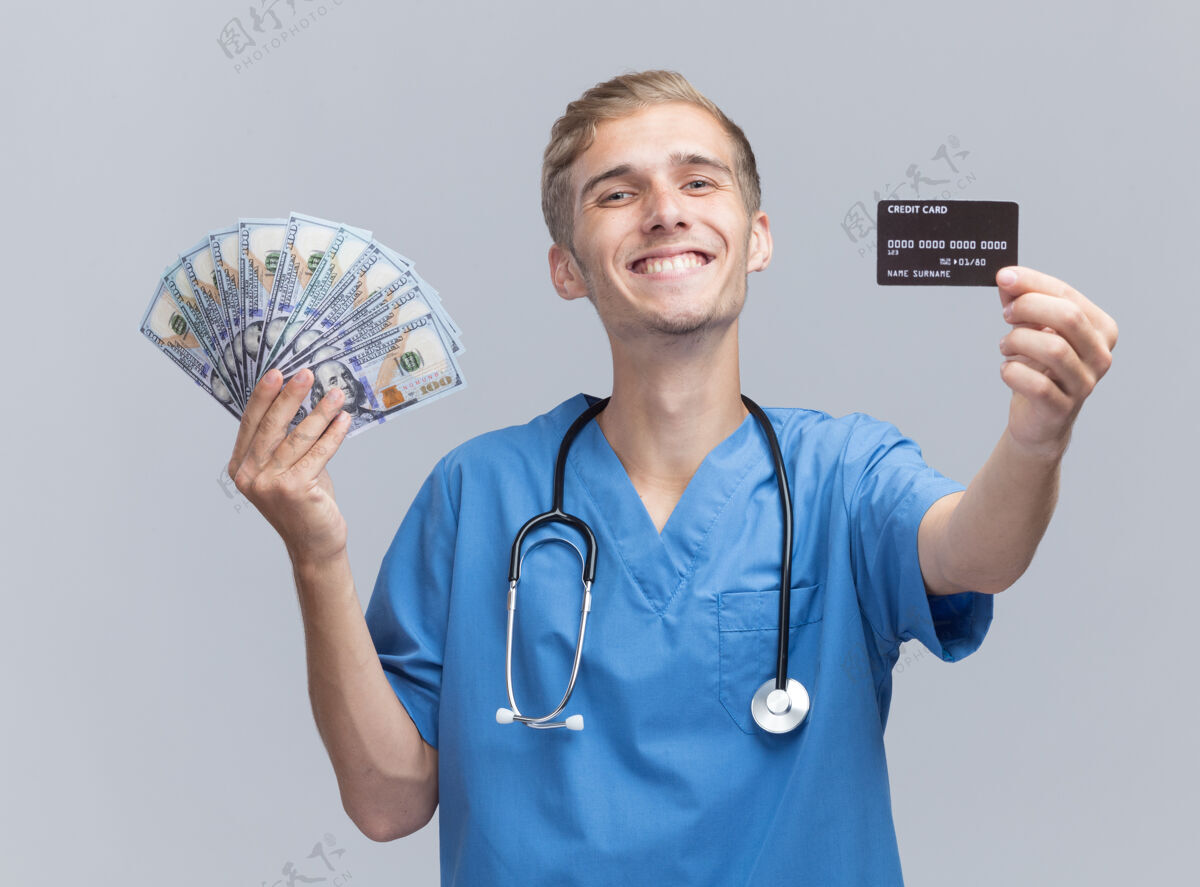 男性微笑的年轻男医生穿着医生制服 手持听诊器 拿着现金和信用卡对着隔离在白墙上的摄像机立场现金制服