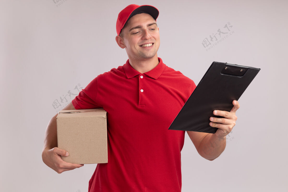 脸面带微笑的年轻送货员穿着制服 拿着帽子盒 看着他手里的剪贴板孤立在白色的墙上站着人衣服