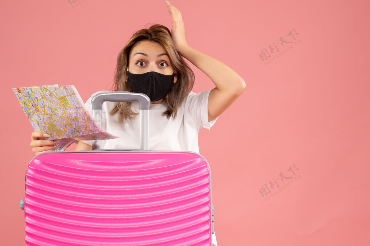 面罩正面图：戴着黑色面具的大眼睛年轻女子拿着地图站在粉色手提箱后面肖像旅游漂亮