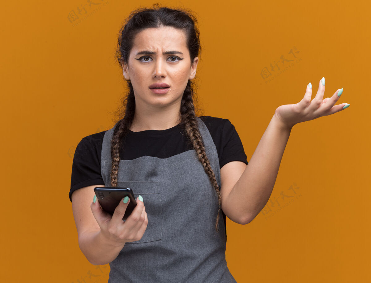 电话迷茫的年轻女理发师穿着制服拿着电话摊开手孤立在橙色的墙上困惑手人