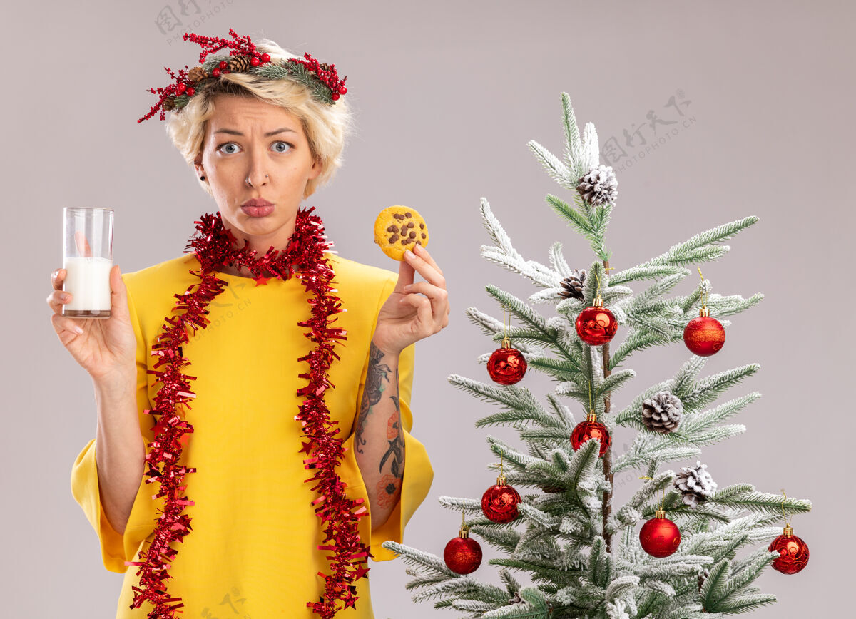 穿困惑的金发女郎头戴圣诞花环 脖子上戴着金属丝花环 站在装饰好的圣诞树旁 手里拿着一杯牛奶和饼干 看上去像是孤零零地站在白色的墙上花环金箔花环