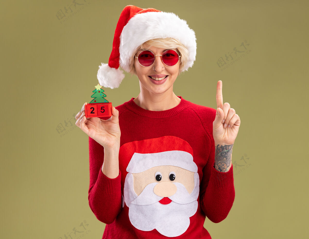 圣诞老人微笑的金发女郎 戴着圣诞帽 戴着圣诞老人的圣诞毛衣 戴着眼镜 手里拿着圣诞树玩具 上面有日期 孤立地指向橄榄绿的墙上 还有复制空间金发圣诞节年轻