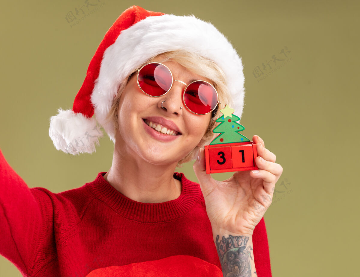视图在橄榄绿的墙上 微笑的金发女郎戴着圣诞帽 穿着圣诞毛衣戴着圣诞眼镜 手里拿着圣诞树玩具 日期看起来很孤立圣诞老人女人举行