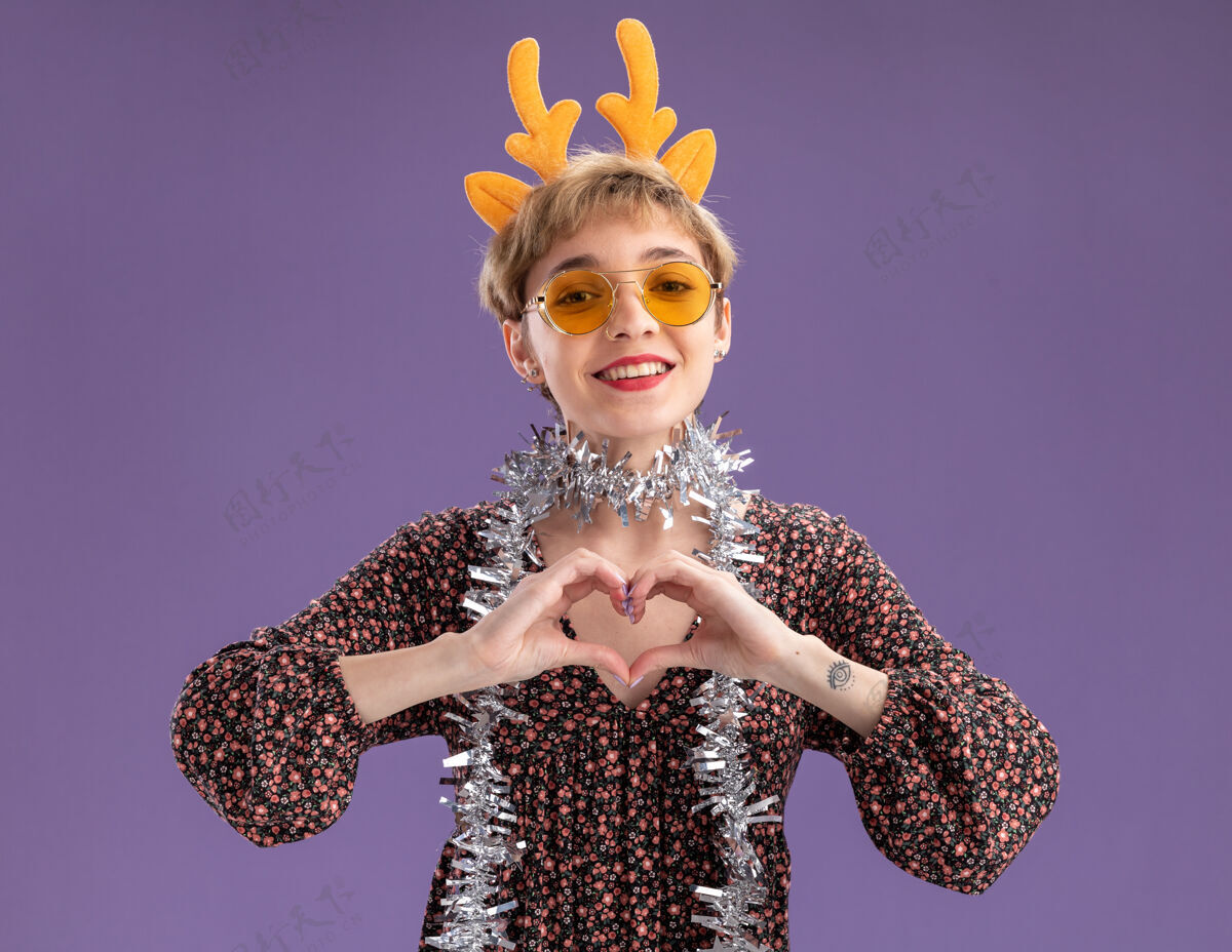 鹿角微笑的年轻漂亮女孩戴着驯鹿鹿角头带 脖子上戴着金箔花环 戴着眼镜 在紫色的墙上做着心形标志眼镜头带金属片