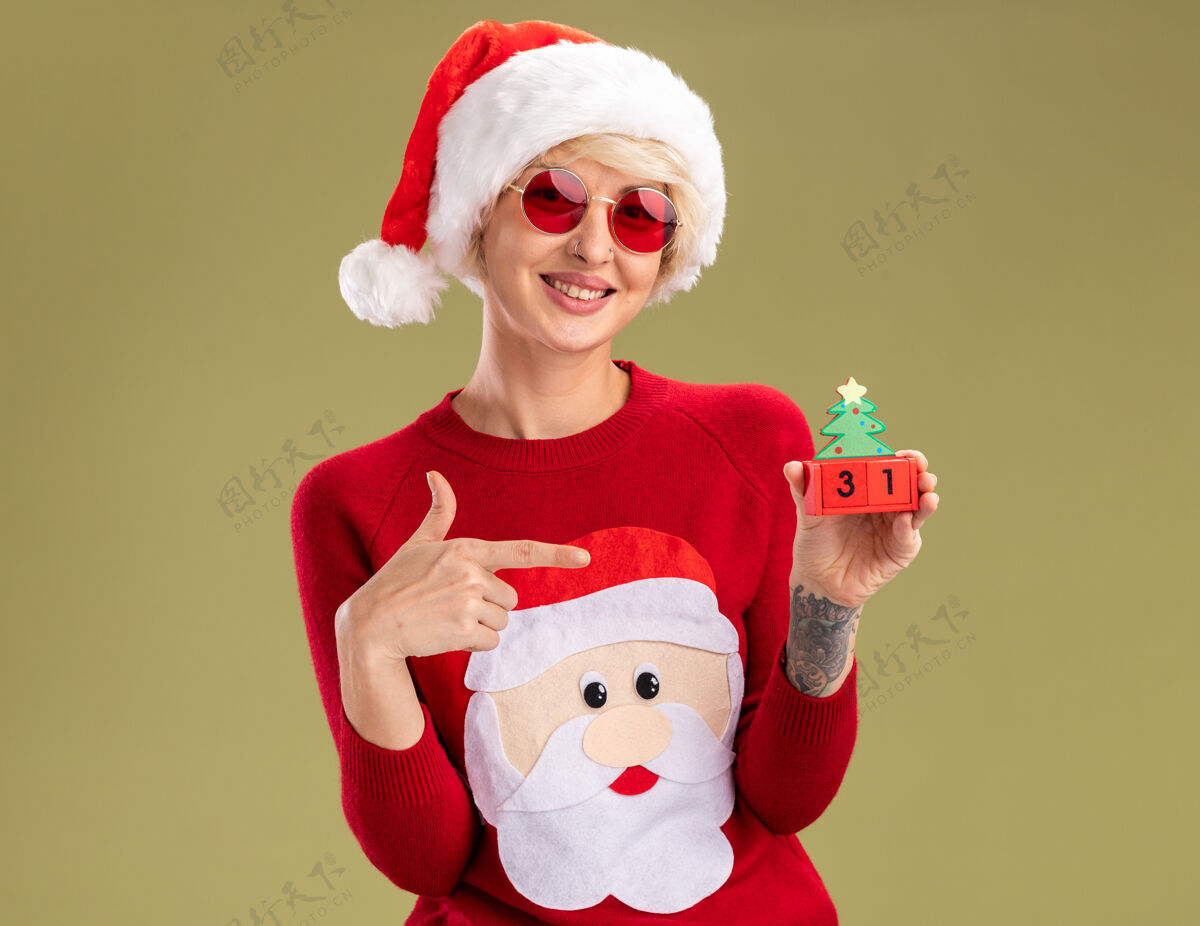 年轻微笑着的金发女郎戴着圣诞帽和圣诞老人的圣诞毛衣 戴着眼镜 拿着并指着橄榄绿墙上的圣诞树玩具 上面的日期看起来很孤立帽子日期毛衣