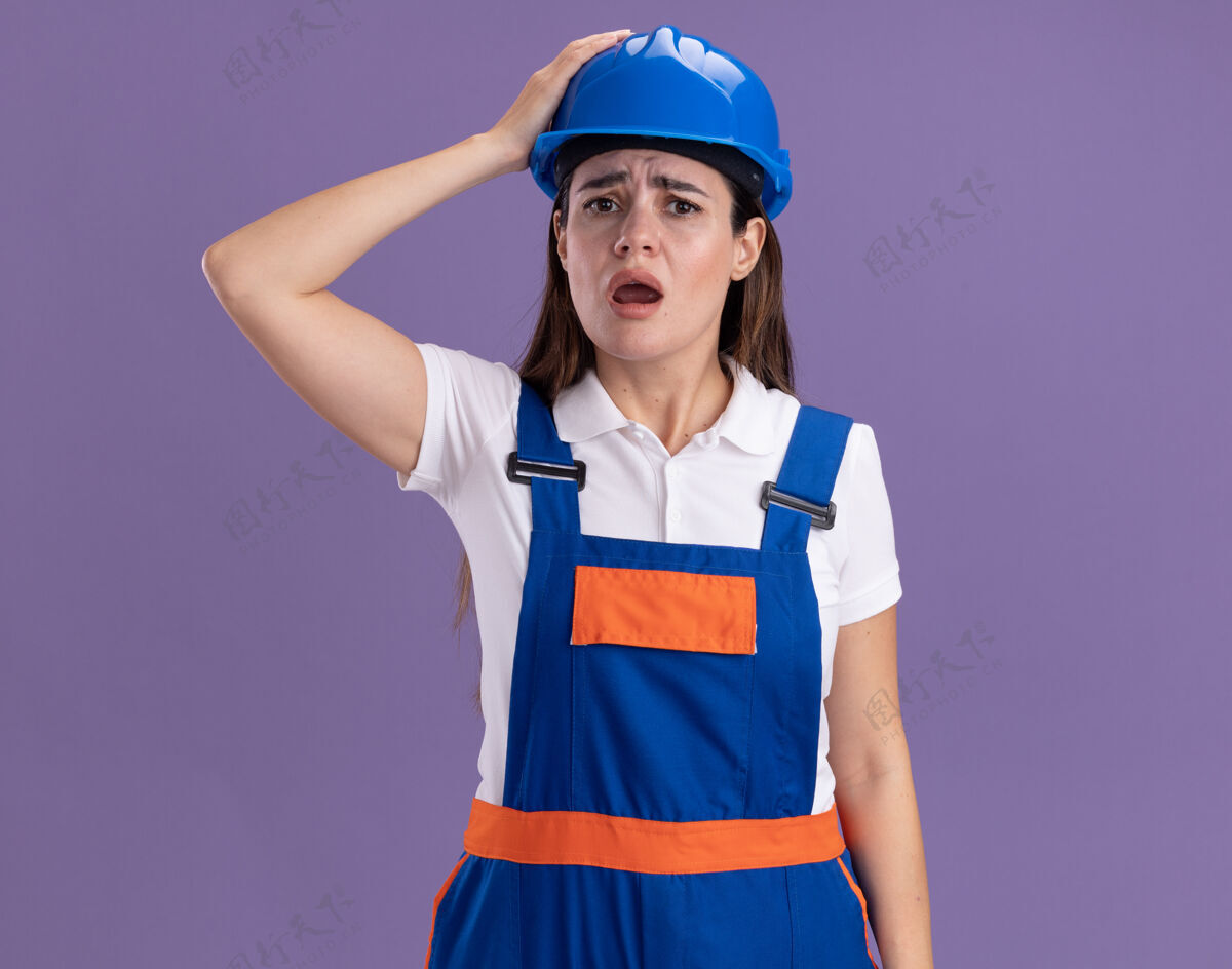 脸一个穿着制服的年轻建筑工人吓得把手放在紫色墙上的头上建设者头立场