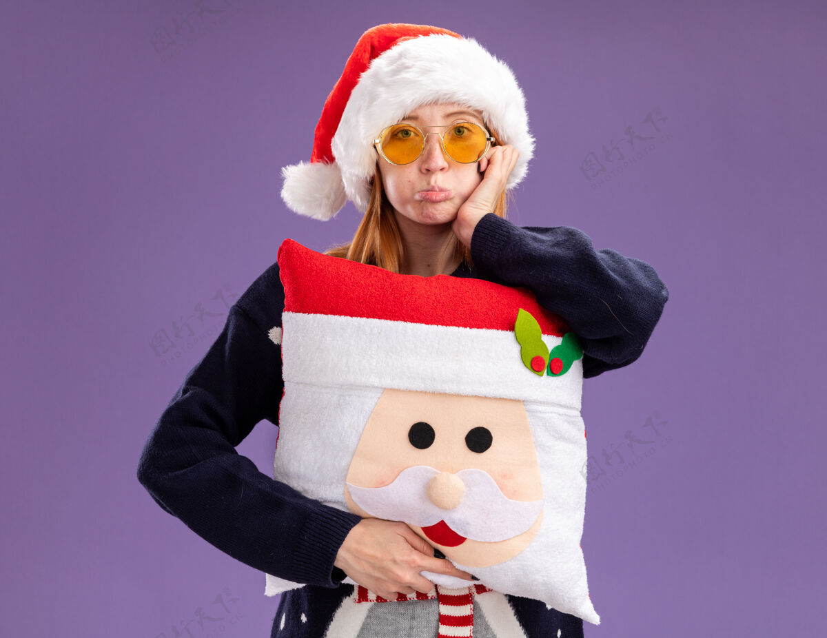 毛衣噘着嘴唇年轻漂亮的女孩穿着圣诞毛衣戴着圣诞帽戴着眼镜拿着圣诞枕头把手放在脸颊上隔离在紫色的墙上手穿枕头