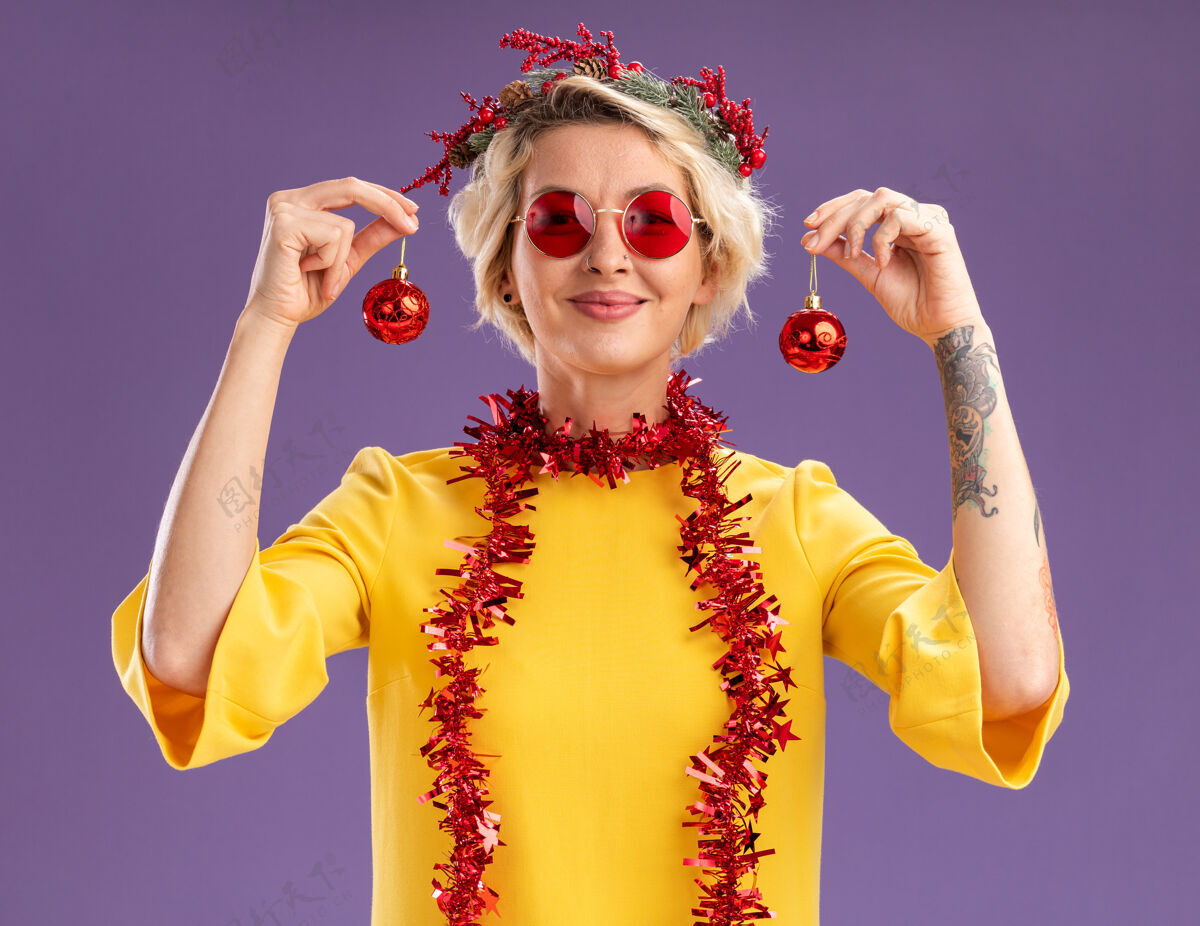饰品戴着圣诞花环 脖子上戴着金箔花环 头上拿着圣诞饰品的年轻金发女郎在紫色的墙上显得孤立无援头近金发