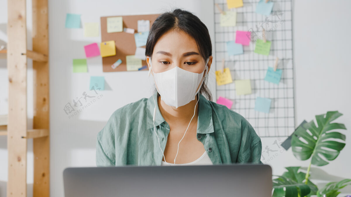 远程戴着医用口罩的亚洲女商人在起居室工作时 用笔记本电脑和同事通过视频通话的方式谈论这个计划流行病耳机办公桌