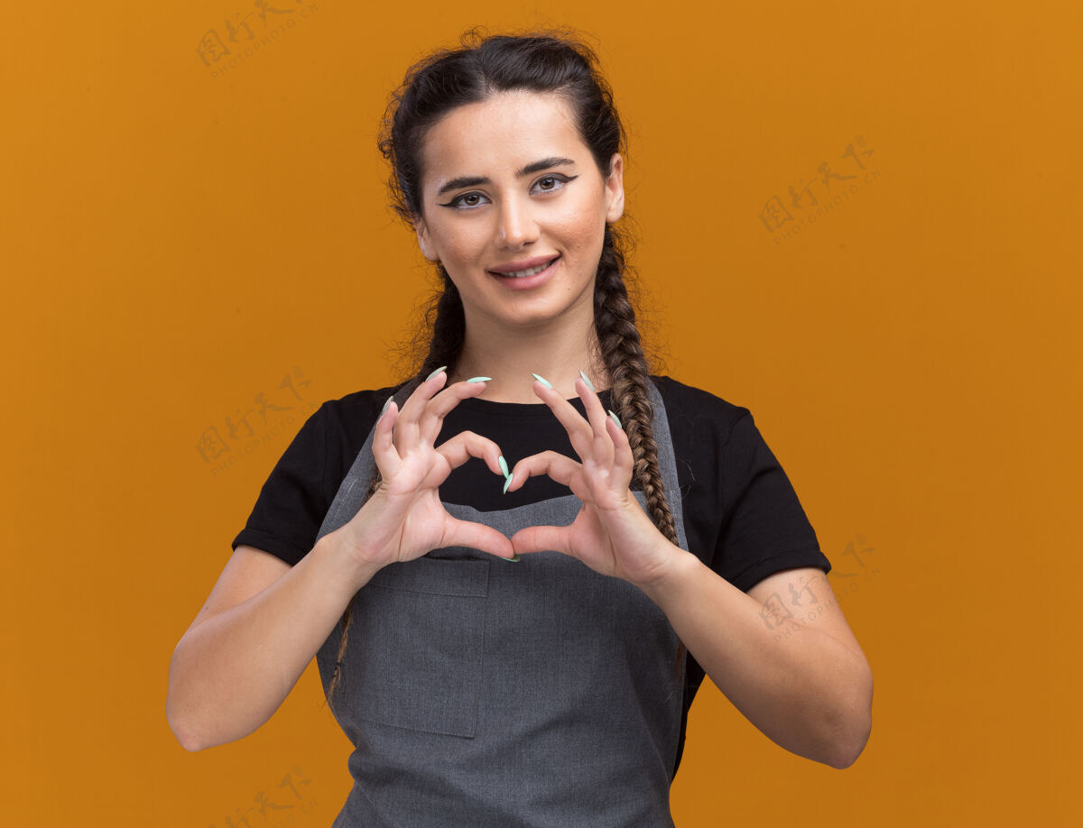 人身着制服的年轻女理发师微笑着 在橙色的墙上展示着孤立的心形脸公民表情