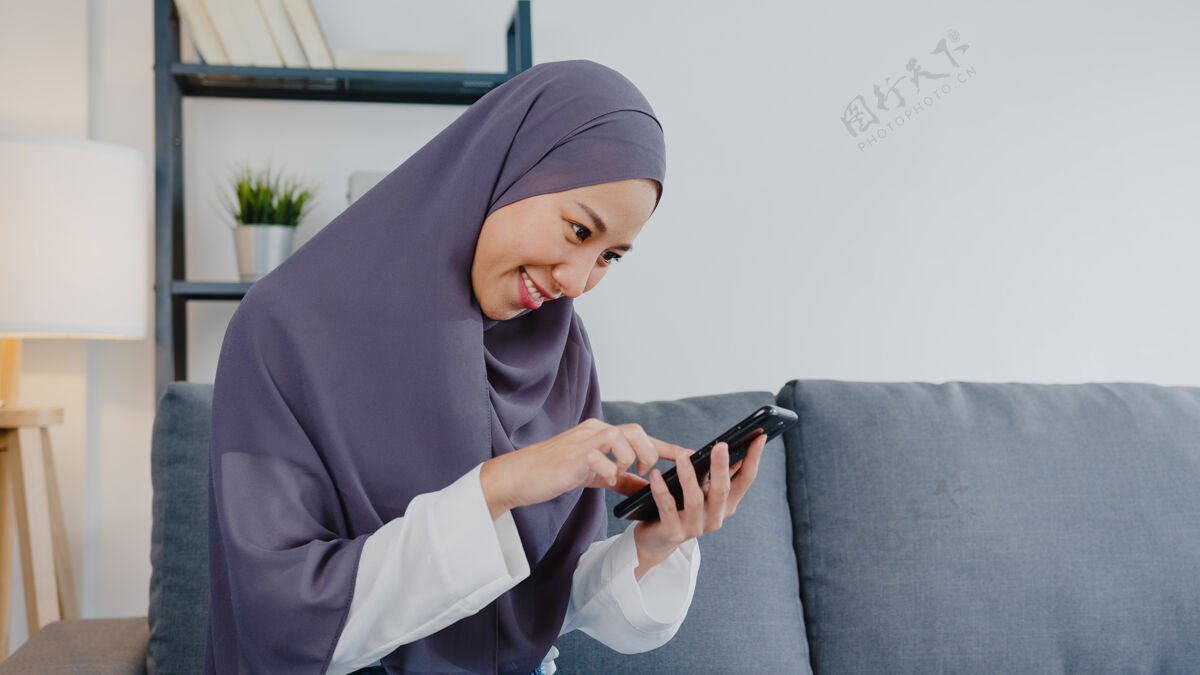 围巾穆斯林女士在家里客厅的沙发上使用智能手机和购买电子商务互联网互联网微笑学生