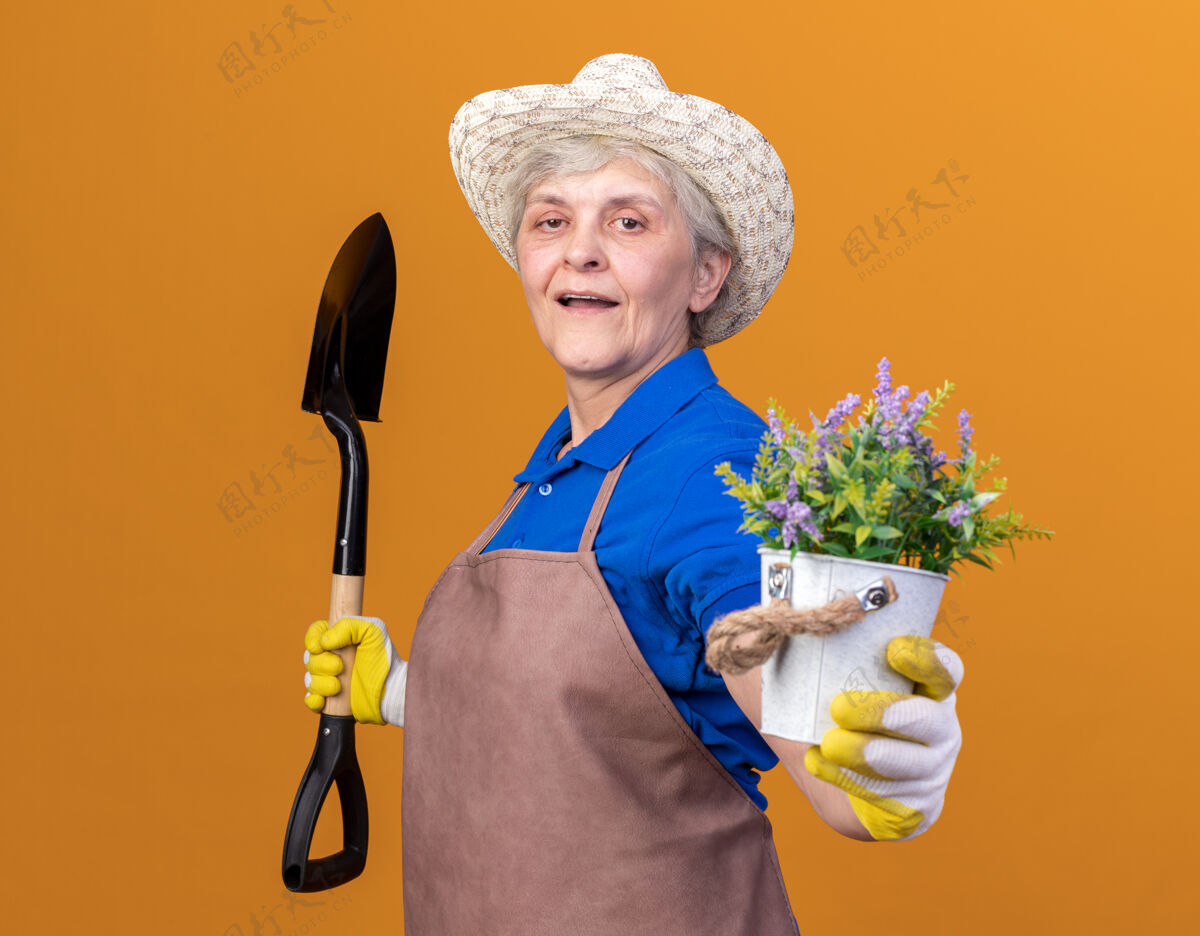 花园自信的上了年纪的女园丁戴着园艺帽和手套站在一边拿着铁锹和花盆侧着站着戴着