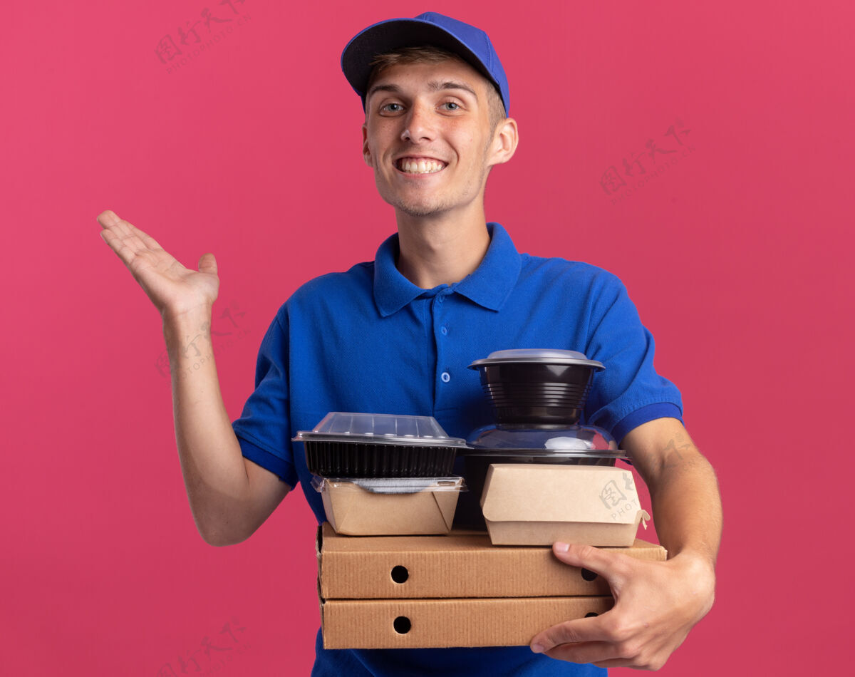 送货微笑着的年轻金发送货员举起手站着 拿着比萨饼盒上的食品容器和包装站粉色提高