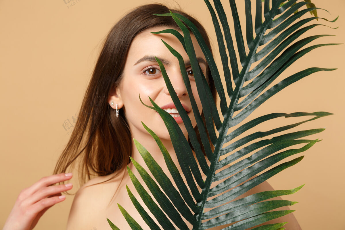 手特写黑发半裸女人完美的皮肤 裸妆和棕榈叶隔离在米色粉彩墙上时尚快乐纯洁