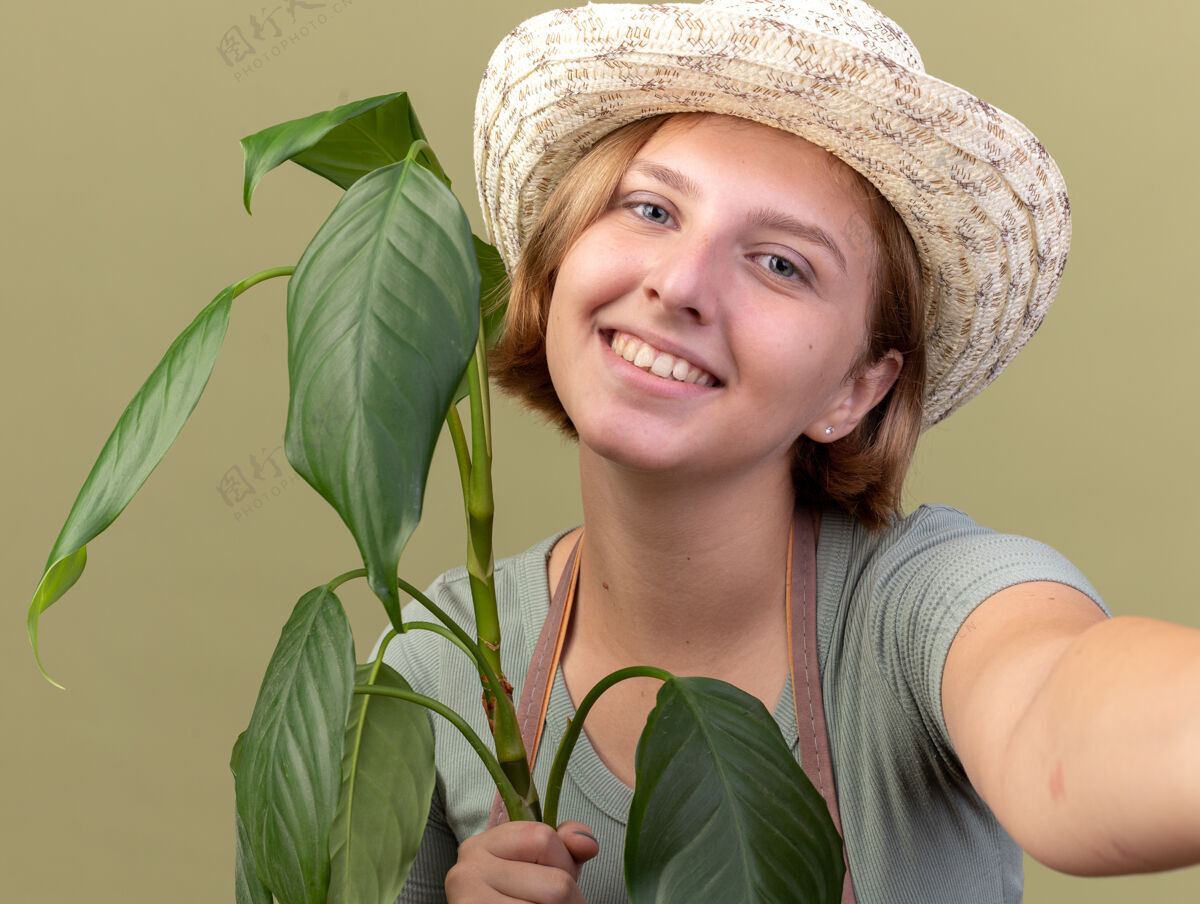 橄榄微笑着的年轻斯拉夫女园丁戴着园艺帽拿着植物假装拿着相机自拍帽子采取微笑