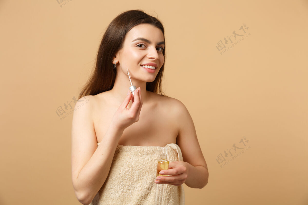 模型特写黑发半裸女人完美的皮肤 裸妆涂抹油从瓶子在脸上隔离在米色粉彩墙健康身体护理浴室