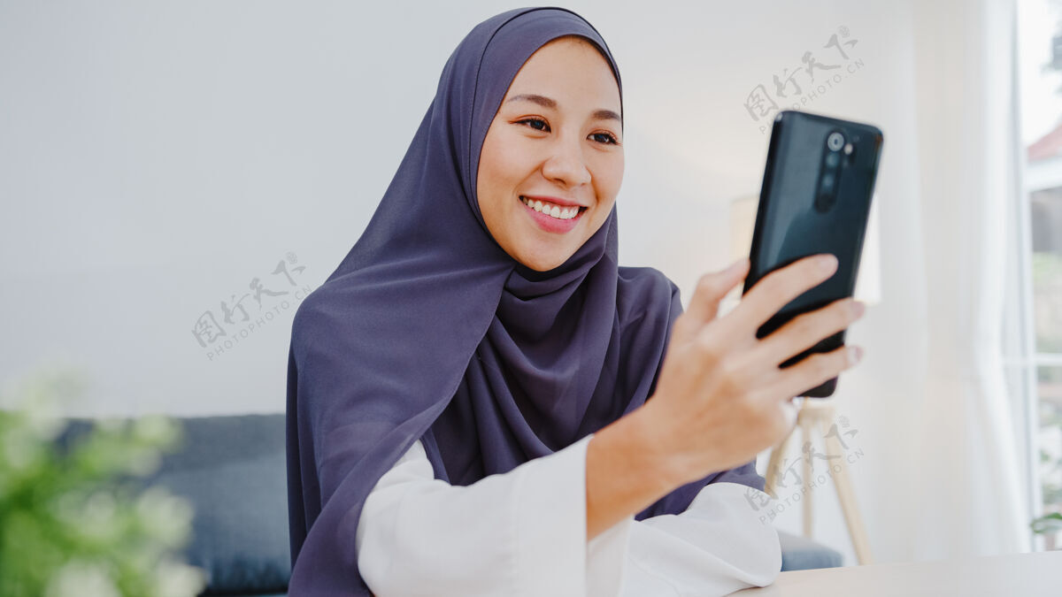 通信年轻的穆斯林女商人使用智能手机通过视频聊天与朋友交谈头脑风暴在线会议 同时在客厅远程工作穆斯林技术视频