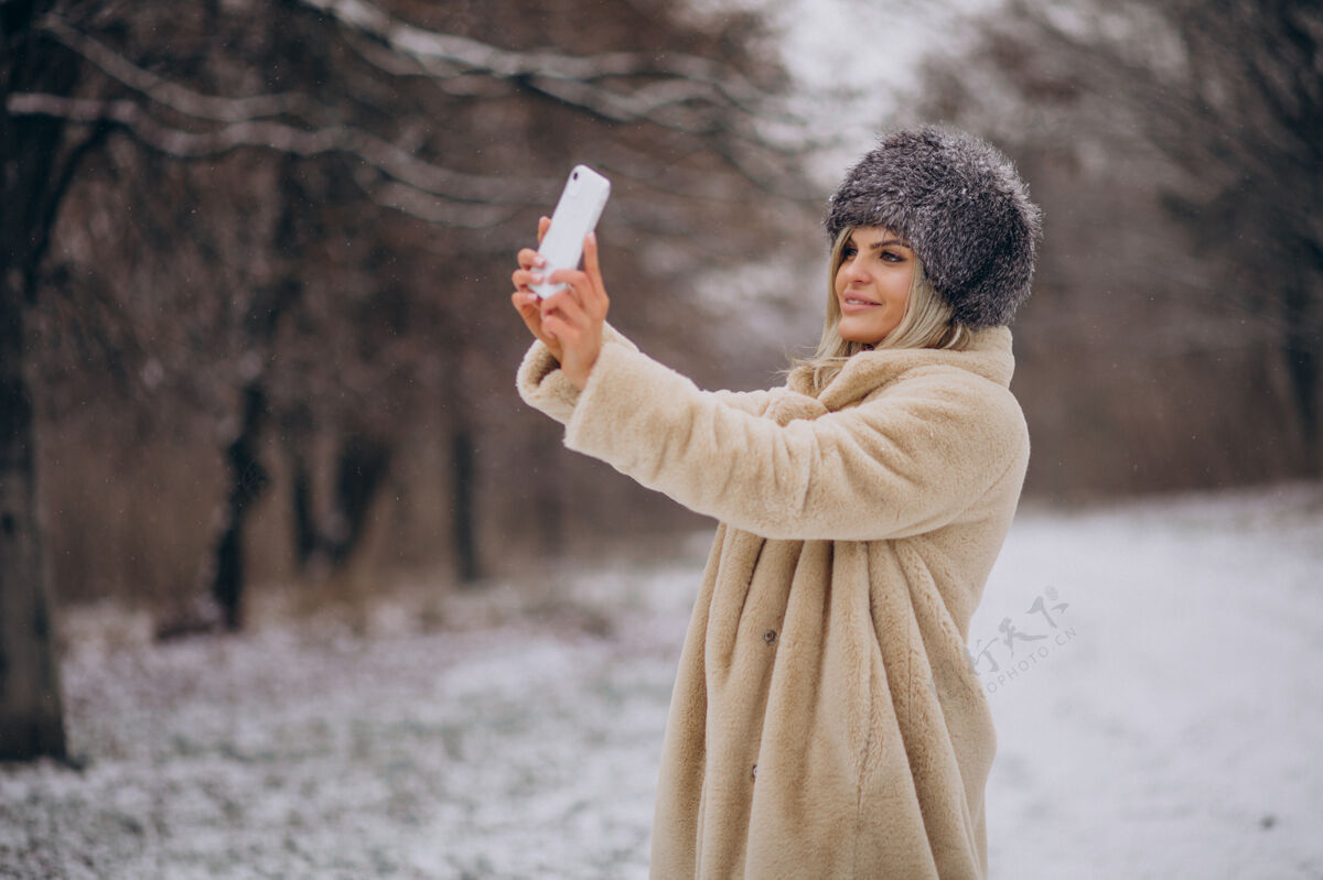 户外穿着冬衣的女人走在满是雪的公园里打电话寒冷树美丽