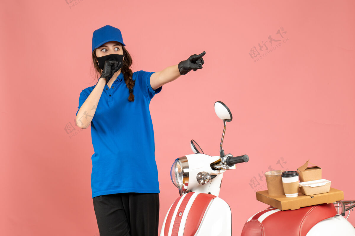 蛋糕俯视图：戴着医用面罩的快递员女孩站在摩托车旁边 摩托车上有咖啡蛋糕 在柔和的桃色背景下向前指背景面具医药
